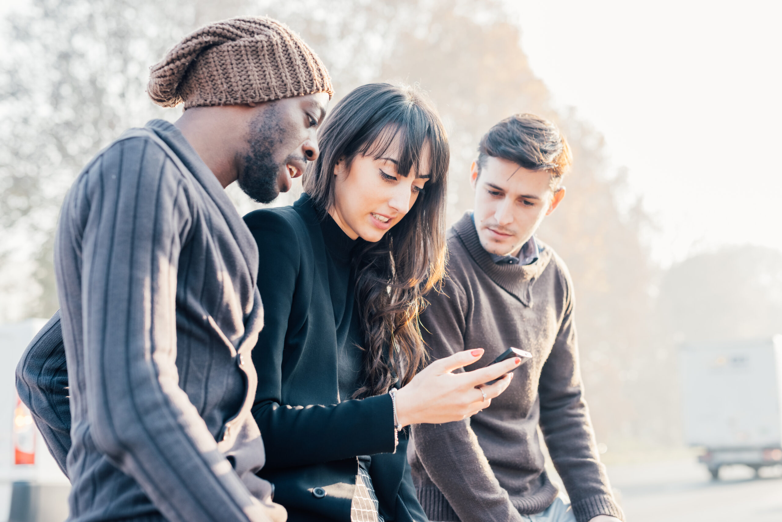Three multiethnic woman and men friends outdoor in city back light, chatting and holding smart phone - friendship, social network, conversation concept