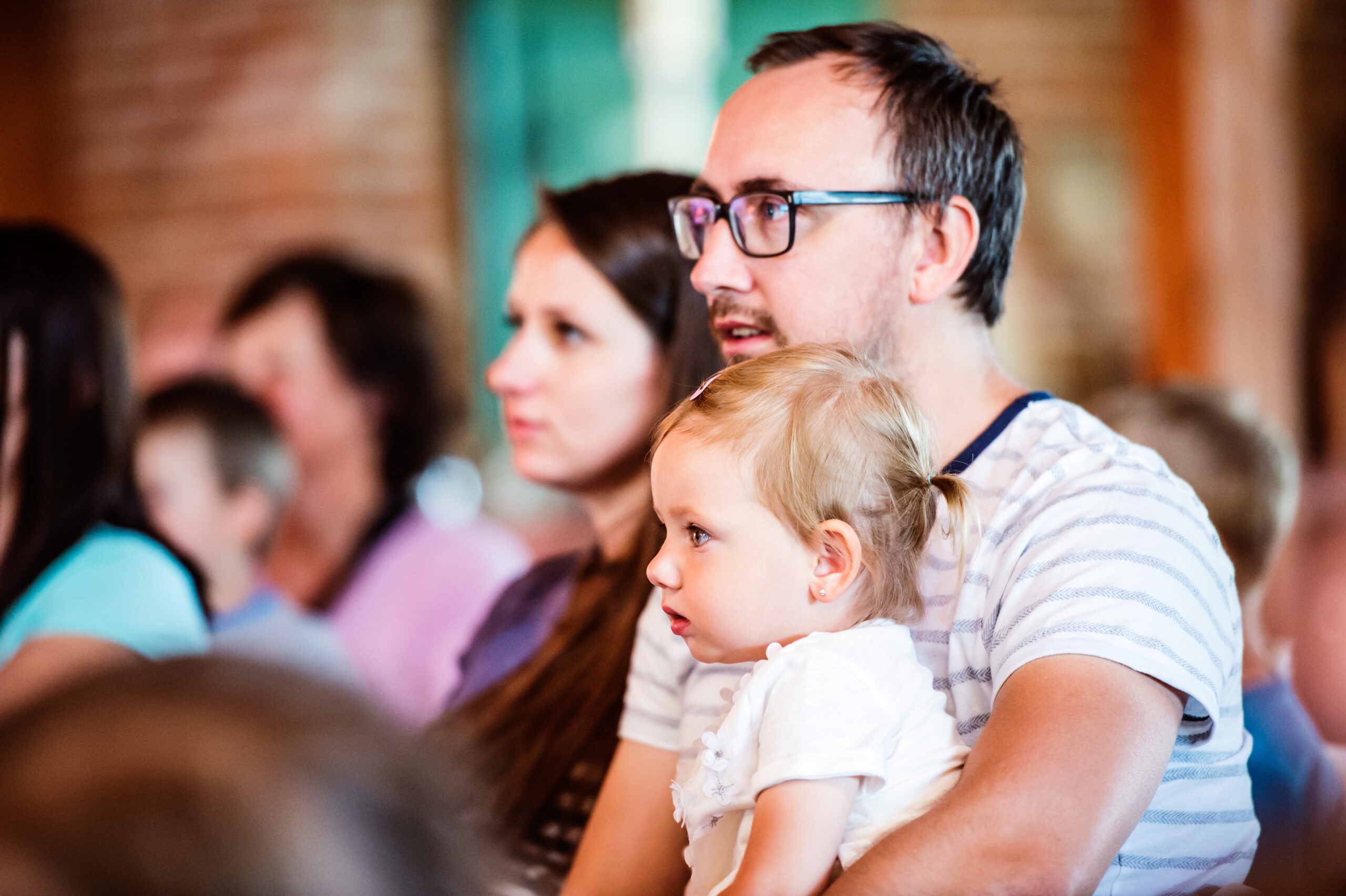 Young family with little daughter sitting outside in a crowd of people, watching show for children